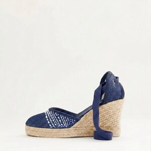 Op de afbeelding: Marineblauwe espadrille sleehak sandaal met een geweven jute zool en een denim bovenwerk. De sandaal heeft een wit en blauw patroon op de teen en een marineblauwe enkelband.