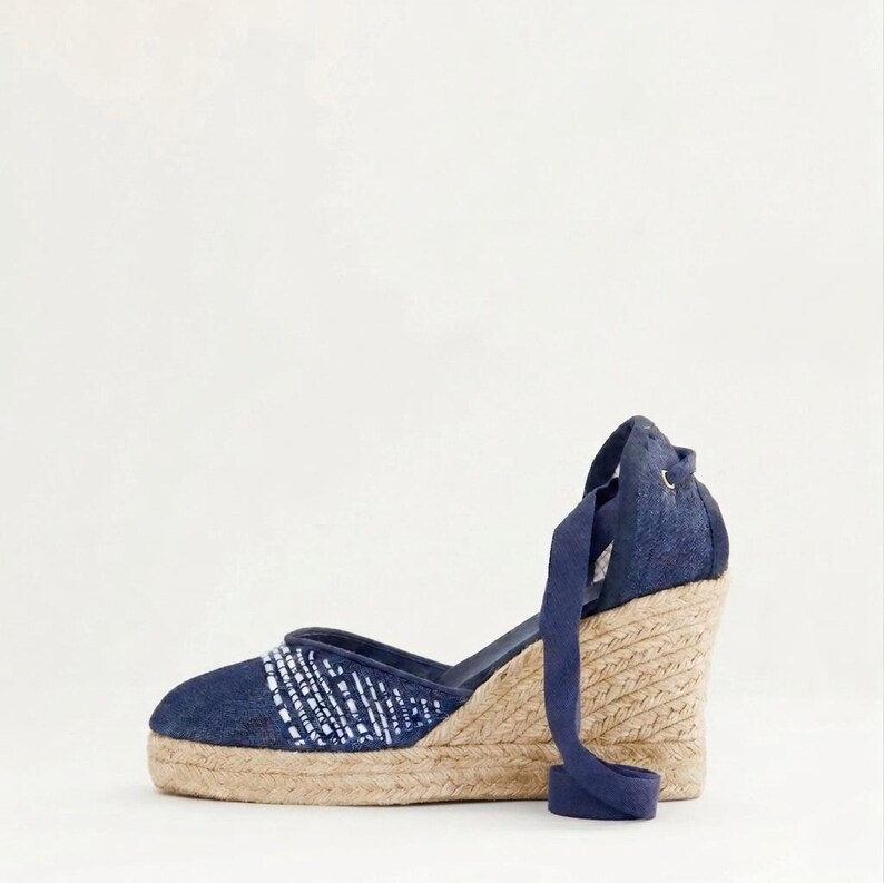 Op de afbeelding: Marineblauwe espadrille sleehak sandaal met een geweven jute zool en een denim bovenwerk. De sandaal heeft een wit en blauw patroon op de teen en een marineblauwe enkelband.