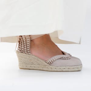May include: Beige espadrille wedge sandals with a woven jute sole and a suede upper. The sandals feature a decorative ruffle detail around the ankle and toe, and a ribbon tie closure. The sandals are shown under a white garment.