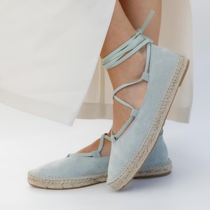May include: Light blue suede espadrille flats with ankle ties. The shoes have a woven jute sole and a rounded toe. The laces wrap around the ankle and tie at the top. The shoes are shown against a white background.
