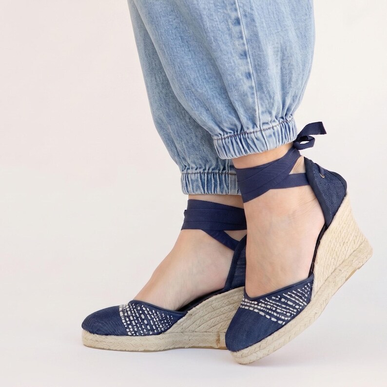 Op de afbeelding: Spijkerstof sleehak espadrilles in blauw met witte borduursels. De schoenen hebben een geweven jute zool en marineblauwe enkelbanden. De sleehak biedt hoogte en ondersteuning, perfect voor een casual, stijlvolle uitstraling.