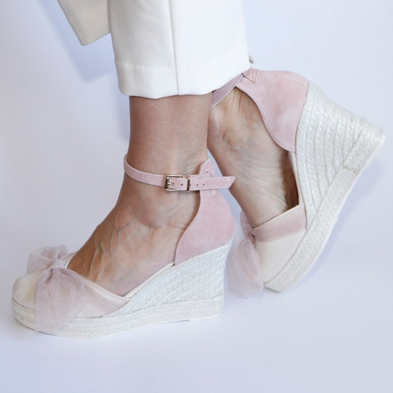 May include: Pink suede wedge sandals with a white platform and a tulle bow detail. The sandals have an ankle strap with a buckle closure.