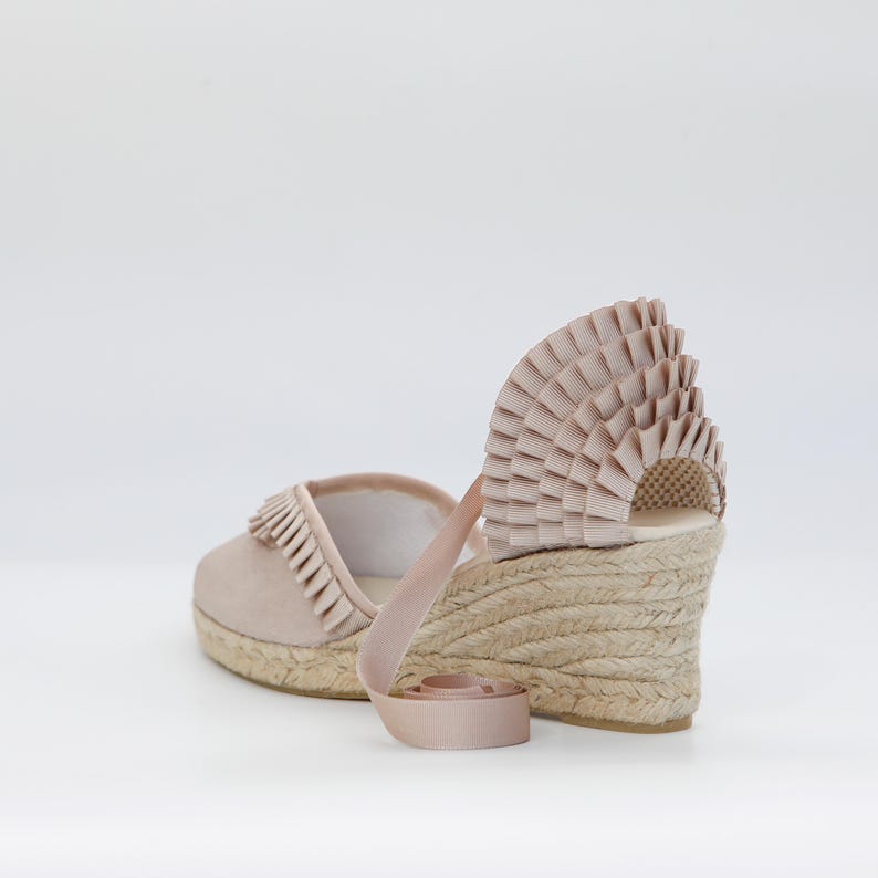 May include: A pair of blush-colored espadrille wedges. The shoes feature a woven jute sole, a suede upper with fringe detail, and a decorative fan-shaped accent on the heel. A ribbon ankle strap adds a feminine touch.