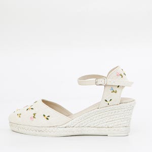 May include: White canvas wedge sandals with an ankle strap and floral embroidery.