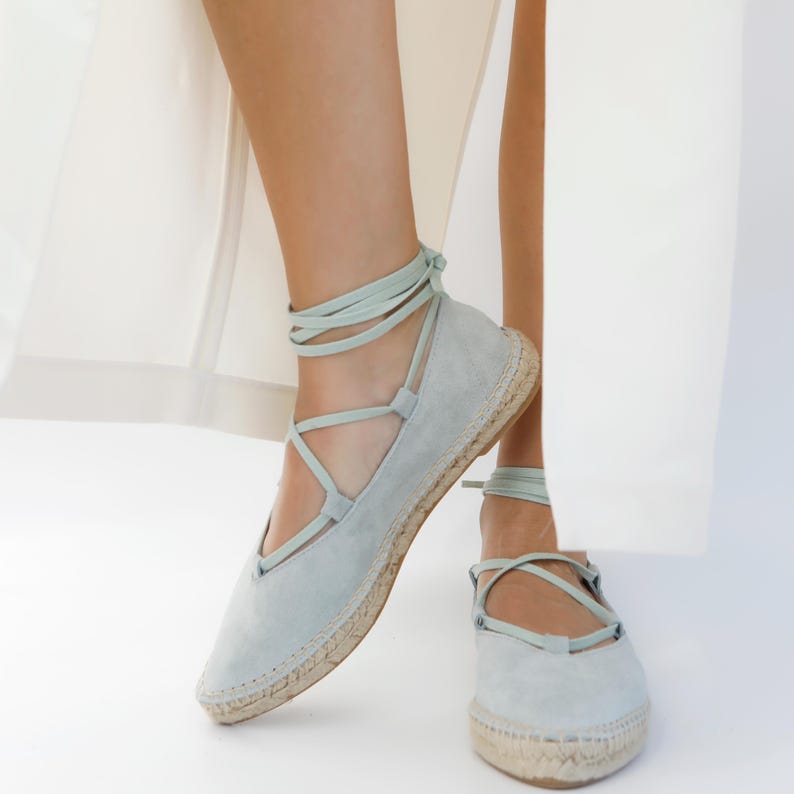 May include: Light blue espadrille flats with ankle ties. The shoes have a suede-like texture and a woven jute sole. The ankle ties are a matching light blue color, wrapping around the ankles. The shoes are shown against a white background.
