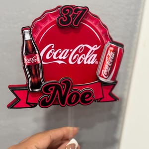 May include: A red and black Coca-Cola themed cake topper with the Coca-Cola logo, a bottle, and a can. The topper also includes the number 37 and the name "Noe".