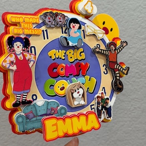 Pode incluir: Um topo de bolo de papel em camadas com um design de mostrador de rel&oacute;gio com o tema The Big Comfy Couch. Inclui personagens, um sof&aacute; azul, o texto Who Made This Big Mess, o n&uacute;mero 4 e o nome Emma em letras amarelas sobre fundo vermelho.