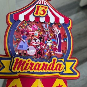 K&ouml;nnte beinhalten: Ein farbenfroher Tortenaufsatz im Zirkus-Design mit der Zahl 13. Der Aufsatz zeigt ein rot-wei&szlig;-blaues Zirkuszelt-Design mit dem Namen "Miranda" in Gelb und Rot. Im Inneren befinden sich Cartoon-Figuren und Konfetti.