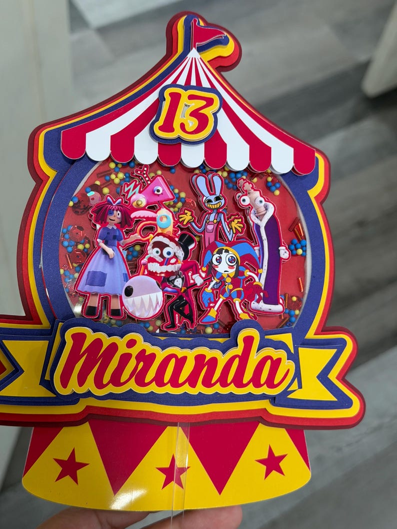 K&ouml;nnte beinhalten: Ein farbenfroher Tortenaufsatz im Zirkus-Design mit der Zahl 13. Der Aufsatz zeigt ein rot-wei&szlig;-blaues Zirkuszelt-Design mit dem Namen "Miranda" in Gelb und Rot. Im Inneren befinden sich Cartoon-Figuren und Konfetti.