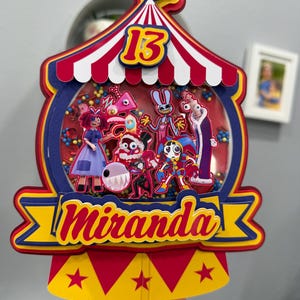 K&ouml;nnte beinhalten: Ein farbenfroher Tortenaufsatz im Zirkus-Design mit der Zahl 13 und dem Namen "Miranda". Der Aufsatz zeigt ein rot-wei&szlig; gestreiftes Zelt-Design mit Cartoon-Figuren in einer transparenten Kuppel, die mit bunten Perlen gef&uuml;llt ist.