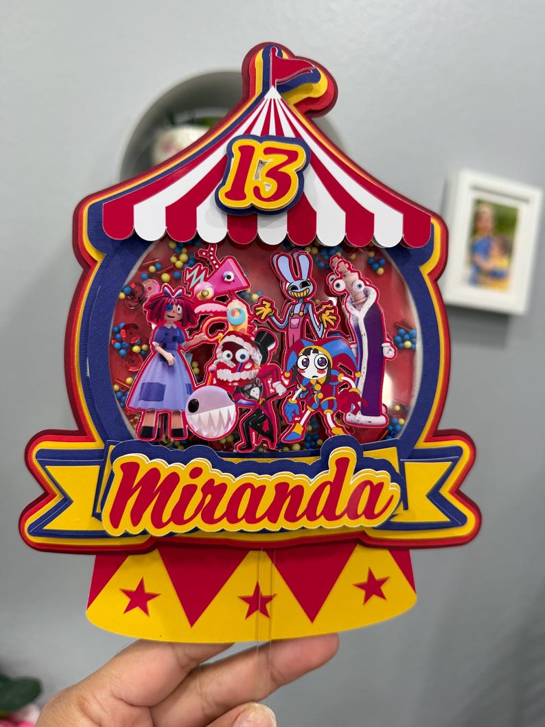 K&ouml;nnte beinhalten: Ein farbenfroher Tortenaufsatz im Zirkus-Design mit der Zahl 13 und dem Namen "Miranda". Der Aufsatz zeigt ein rot-wei&szlig; gestreiftes Zelt-Design mit Cartoon-Figuren in einer transparenten Kuppel, die mit bunten Perlen gef&uuml;llt ist.