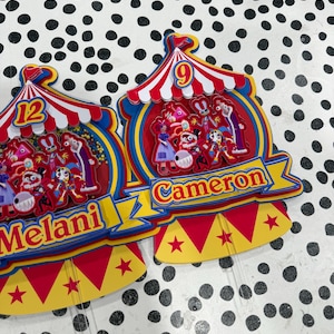 K&ouml;nnte beinhalten: Zwei farbenfrohe Zirkus-Themen-Kuchenaufs&auml;tze. Einer hat die Zahl 12 und den Namen "Melani". Der andere hat die Zahl 9 und den Namen "Cameron". Beide Aufs&auml;tze haben rote, gelbe und blaue Farben.
