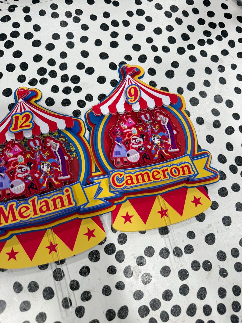 K&ouml;nnte beinhalten: Zwei farbenfrohe Zirkus-Themen-Kuchenaufs&auml;tze. Einer hat die Zahl 12 und den Namen "Melani". Der andere hat die Zahl 9 und den Namen "Cameron". Beide Aufs&auml;tze haben rote, gelbe und blaue Farben.