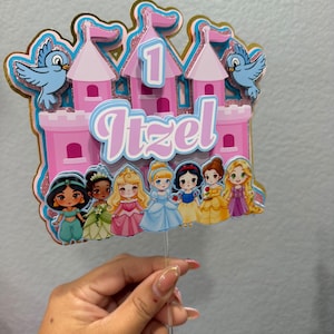 Pode incluir: Um topo de bolo multicamadas com um castelo rosa, o número 1, o nome Itzel e sete princesas de desenho animado. O design inclui dois pássaros azuis nas laterais, detalhes com glitter e uma haste de plástico transparente para fixação.