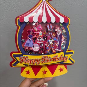 K&ouml;nnte beinhalten: Ein farbenfroher Geburtstagskuchenaufsatz im Zirkus-Design. Der Aufsatz zeigt ein rot-wei&szlig; gestreiftes Zelt-Design mit verschiedenen Zeichentrickfiguren im Inneren. Die Worte "Happy Birthday" sind auf einem gelben Banner zu sehen.