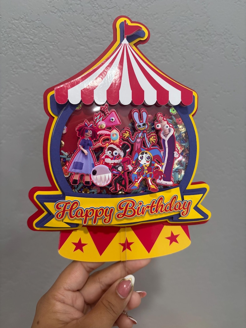 K&ouml;nnte beinhalten: Ein farbenfroher Geburtstagskuchenaufsatz im Zirkus-Design. Der Aufsatz zeigt ein rot-wei&szlig; gestreiftes Zelt-Design mit verschiedenen Zeichentrickfiguren im Inneren. Die Worte "Happy Birthday" sind auf einem gelben Banner zu sehen.
