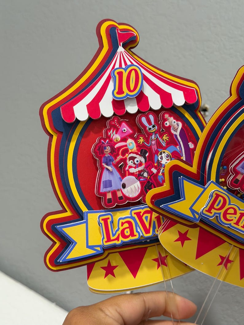 K&ouml;nnte beinhalten: Ein farbenfroher Tortenaufsatz im Zirkus-Design mit der Zahl 10. Das Design zeigt ein rot-wei&szlig; gestreiftes Zelt, Cartoon-Figuren und den Namen "Lavi" auf einem Banner. Der Aufsatz ist mit roten, gelben und blauen Akzenten versehen.