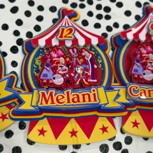 K&ouml;nnte beinhalten: Bunte Zirkus-Themen-Kuchenaufs&auml;tze mit dem Namen "Melani" auf einem gelben Banner. Die Aufs&auml;tze zeigen ein rot-wei&szlig; gestreiftes Zelt-Design mit der Zahl 12 und verschiedenen Cartoon-Figuren. Die Basis hat rote, gelbe und blaue Akzente.
