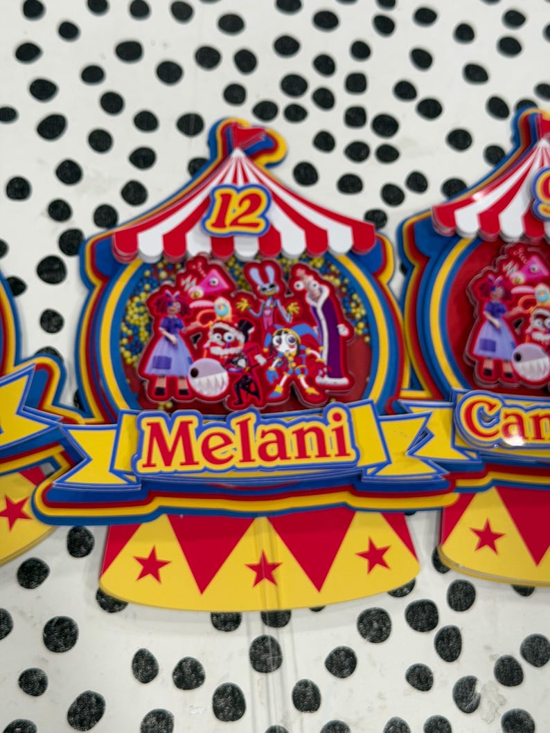 K&ouml;nnte beinhalten: Bunte Zirkus-Themen-Kuchenaufs&auml;tze mit dem Namen "Melani" auf einem gelben Banner. Die Aufs&auml;tze zeigen ein rot-wei&szlig; gestreiftes Zelt-Design mit der Zahl 12 und verschiedenen Cartoon-Figuren. Die Basis hat rote, gelbe und blaue Akzente.