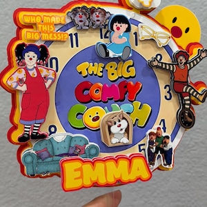 Pode incluir: Um topo de bolo em camadas com design de mostrador de rel&oacute;gio e o texto "The Big Comfy Couch" e "Who Made This Big Mess!?". Inclui recortes de personagens, um sof&aacute; azul, &oacute;culos amarelos e o nome "Emma" em amarelo, com um n&uacute;mero 4 no topo.