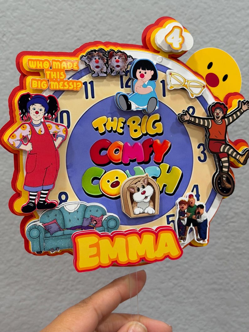 Pode incluir: Um topo de bolo em camadas com design de mostrador de rel&oacute;gio e o texto "The Big Comfy Couch" e "Who Made This Big Mess!?". Inclui recortes de personagens, um sof&aacute; azul, &oacute;culos amarelos e o nome "Emma" em amarelo, com um n&uacute;mero 4 no topo.