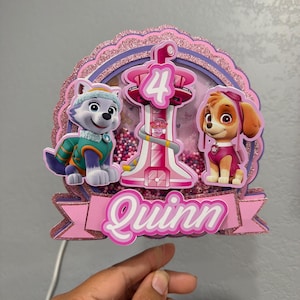 Pode incluir: Um topo de bolo em camadas, brilhante em rosa e roxo, com as personagens Everest e Skye da Patrulha Pata, uma torre central com o número 4 e uma faixa na parte inferior com o nome Quinn impresso em branco sobre um fundo rosa.