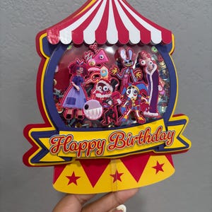 K&ouml;nnte beinhalten: Ein farbenfroher Geburtstagskuchenaufsatz in Form eines Zirkuszeltes. Der Aufsatz zeigt verschiedene Cartoon-Figuren in einem transparenten Fenster, mit dem Schriftzug "Happy Birthday" auf einem Banner. Rot, Gelb, Blau und Wei&szlig; sind die Hauptfarben.