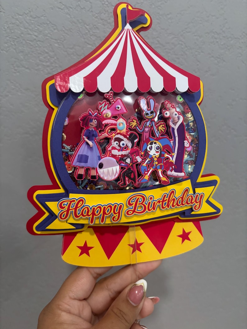K&ouml;nnte beinhalten: Ein farbenfroher Geburtstagskuchenaufsatz in Form eines Zirkuszeltes. Der Aufsatz zeigt verschiedene Cartoon-Figuren in einem transparenten Fenster, mit dem Schriftzug "Happy Birthday" auf einem Banner. Rot, Gelb, Blau und Wei&szlig; sind die Hauptfarben.
