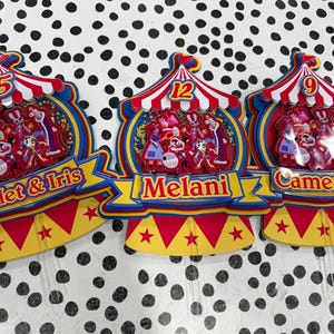 K&ouml;nnte beinhalten: Bunte Geburtstagskuchenaufs&auml;tze im Zirkus-Design. Jeder Aufsatz hat ein rot-wei&szlig; gestreiftes Zeltmuster, eine Zahl und ein Namensbanner. Die Aufs&auml;tze sind mit Zeichentrickfiguren und Sternen verziert.
