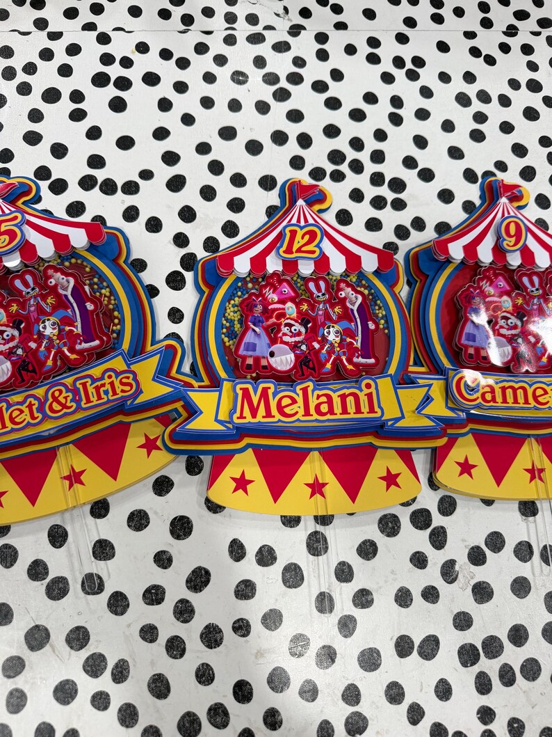 K&ouml;nnte beinhalten: Bunte Geburtstagskuchenaufs&auml;tze im Zirkus-Design. Jeder Aufsatz hat ein rot-wei&szlig; gestreiftes Zeltmuster, eine Zahl und ein Namensbanner. Die Aufs&auml;tze sind mit Zeichentrickfiguren und Sternen verziert.
