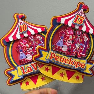 K&ouml;nnte beinhalten: Zwei farbenfrohe Tortenaufs&auml;tze im Zirkus-Design. Jeder hat ein rot-wei&szlig; gestreiftes Zelt, ein Banner mit einem Namen und einer Zahl. Einer sagt "Lavy" mit der Nummer 10, der andere sagt "Penelope" mit der Nummer 13.