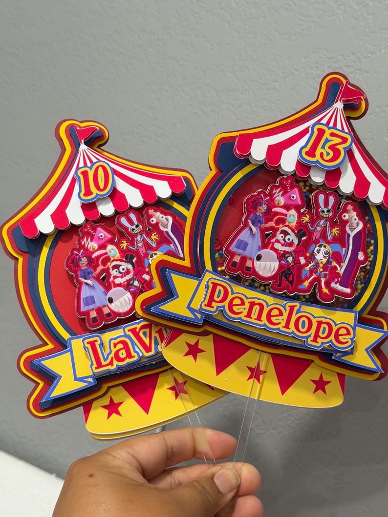 K&ouml;nnte beinhalten: Zwei farbenfrohe Tortenaufs&auml;tze im Zirkus-Design. Jeder hat ein rot-wei&szlig; gestreiftes Zelt, ein Banner mit einem Namen und einer Zahl. Einer sagt "Lavy" mit der Nummer 10, der andere sagt "Penelope" mit der Nummer 13.
