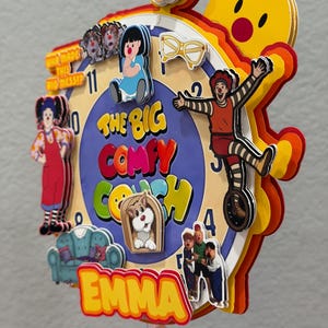Pode incluir: Um topo de bolo de papel multicamadas com um design de mostrador de rel&oacute;gio e personagens de The Big Comfy Couch, incluindo Loonette e Molly. O texto diz The Big Comfy Couch, Emma e Who Made This Big Mess. Inclui um n&uacute;mero 4 no topo.