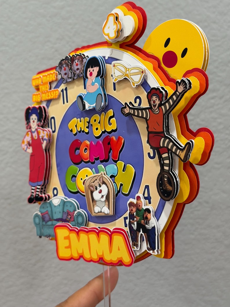 Pode incluir: Um topo de bolo de papel multicamadas com um design de mostrador de rel&oacute;gio e personagens de The Big Comfy Couch, incluindo Loonette e Molly. O texto diz The Big Comfy Couch, Emma e Who Made This Big Mess. Inclui um n&uacute;mero 4 no topo.