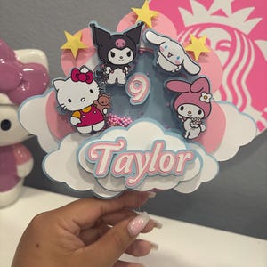Pode incluir: Um topo de bolo em camadas com personagens de desenhos animados, incluindo Hello Kitty, Kuromi e My Melody, com o nome "Taylor" em escrita rosa. O design incorpora nuvens, estrelas e o número "9".