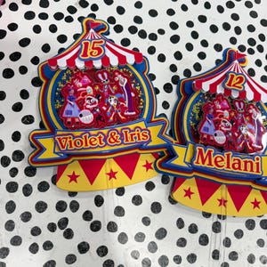 K&ouml;nnte beinhalten: Zwei farbenfrohe Tortenaufs&auml;tze im Zirkus-Design. Einer hat die Zahl 15, den Text "Violet & Iris" und ein Zirkuszelt-Design. Der andere hat die Zahl 12 und den Namen "Melani". Beide haben Zeichentrickfiguren.