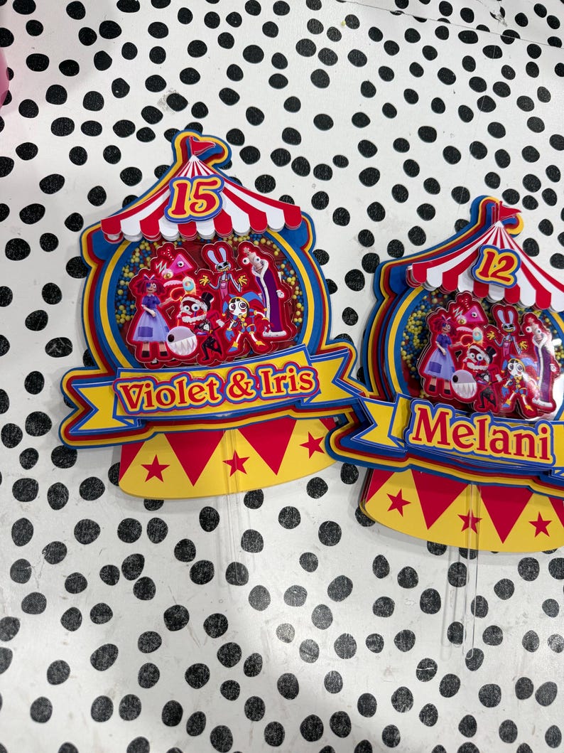 K&ouml;nnte beinhalten: Zwei farbenfrohe Tortenaufs&auml;tze im Zirkus-Design. Einer hat die Zahl 15, den Text "Violet & Iris" und ein Zirkuszelt-Design. Der andere hat die Zahl 12 und den Namen "Melani". Beide haben Zeichentrickfiguren.