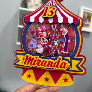 K&ouml;nnte beinhalten: Ein farbenfroher Kuchenaufsatz im Zirkus-Design mit der Zahl 13 und dem Namen "Miranda". Der Aufsatz zeigt ein rot-wei&szlig; gestreiftes Zelt-Design mit Cartoon-Figuren im Inneren. Das Banner ist gelb und blau.