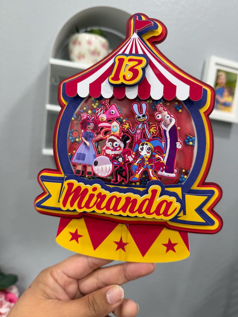 K&ouml;nnte beinhalten: Ein farbenfroher Kuchenaufsatz im Zirkus-Design mit der Zahl 13 und dem Namen "Miranda". Der Aufsatz zeigt ein rot-wei&szlig; gestreiftes Zelt-Design mit Cartoon-Figuren im Inneren. Das Banner ist gelb und blau.
