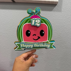 May include: A layered paper cake topper featuring a cartoon watermelon character with a pink face, black eyes, and a smile. The number 12 is displayed at the top, and a green banner at the bottom contains the white text Happy Birthday.
