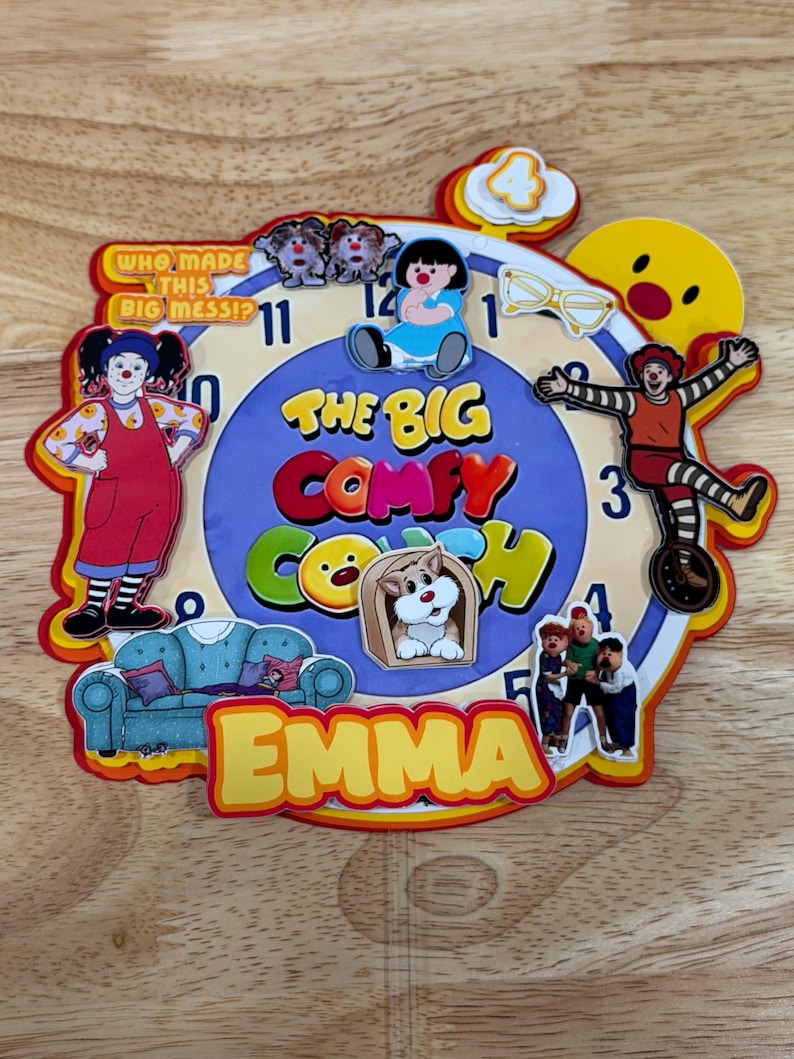 Pode incluir: Um rel&oacute;gio de parede circular com o tema The Big Comfy Couch, mostrador azul, n&uacute;meros pretos e recortes de personagens coloridos. O texto diz Who Made This Big Mess, The Big Comfy Couch e o nome Emma em letras amarelas na parte inferior.