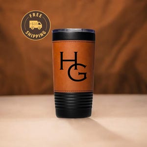 Può includere: Un bicchiere in acciaio inossidabile rivestito in pelle marrone con un coperchio nero e un monogramma delle lettere "HG" in nero.