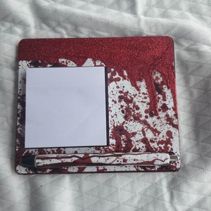 May include: A red and white desk pad with a blood splatter design. The pad has a white sticky note and a silver pen.