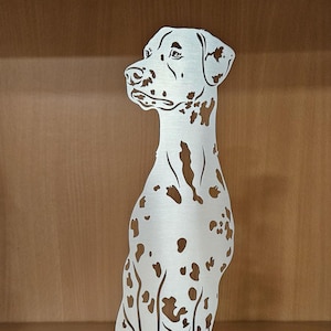 Könnte beinhalten: Metallskulptur eines sitzenden Dalmatinerhundes. Der Hund ist weiß mit braunen Flecken, und die Skulptur hat eine gebürstete Metalloberfläche. Der Hund sitzt auf einem rechteckigen Sockel.