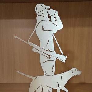 May include: A silver metal sculpture of a hunter with a dog. The hunter is holding binoculars and a rifle. The dog is standing next to the hunter and is looking to the right. The sculpture is set on a base with grass.