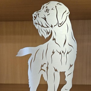 Op de afbeelding: Een zilveren metalen silhouet van een hond die in het gras staat. De hond kijkt naar links en heeft zijn hoofd opgeheven. De hond is een draadhaarras met een lange staart.