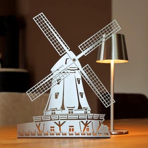 Könnte beinhalten: Eine weiße Metallwindmühlen-Skulptur mit detailliertem Ausschnitt-Design. Die Windmühle hat vier Flügel, einen detaillierten Körper und einen Sockel mit ausgeschnittenem Gras und Häusern. Neben der Windmühle steht eine silberne Lampe mit konischem Schirm.