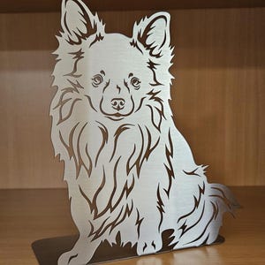 以下が含まれることがあります： 頭が少し横に傾けられた状態で座っている、長毛のチワワの銀色の金属彫刻。彫刻は毛皮と顔の特徴で詳細に作られています。