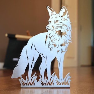 Könnte beinhalten: Eine silberne Metallfuchs-Skulptur, die im Gras steht. Der Fuchs ist in einem detaillierten, ausgeschnittenen Design dargestellt, mit zur Seite gedrehtem Kopf. Die Skulptur steht auf einer Holzoberfläche.