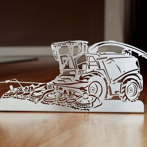 Può includere: Una scultura in metallo color argento di una trinciatrice CLAAS. Il design dettagliato a ritaglio raffigura la cabina, le ruote e la testata di taglio della macchina. La scultura è esposta su una superficie in legno.